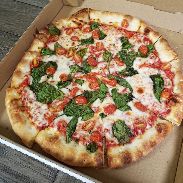 A whole Pizza 72 pizza in a cardboard box, topped with melted cheese, spinach, and cherry tomatoes.